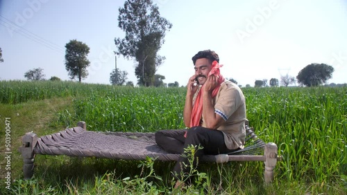 A handsome bearded farmer talking on his smartphone while relaxing on a Chaarpai - Green wheat field. An Indian villager wiping the sweat on his forehead while using an Angocha - summertime  agricu... photo