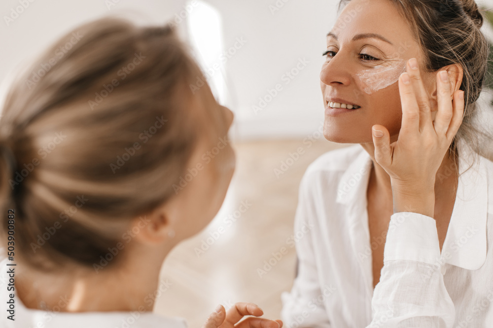 Fair-skinned girl applies moisturizer to face of adult woman on white background. Blond woman with flawless skin in casual shirt. Skin care and beauty concept Fair-skinned girl applies moisturizer to face of adult woman on white background. Blond woman with flawless skin in casual shirt. Skin care and beauty concept
