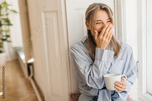 Cheerful caucasian young girl drinks coffee and laughs, covering her face with hand, sitting by window in light room. Blonde in good mood wears shirt. Emotion concept