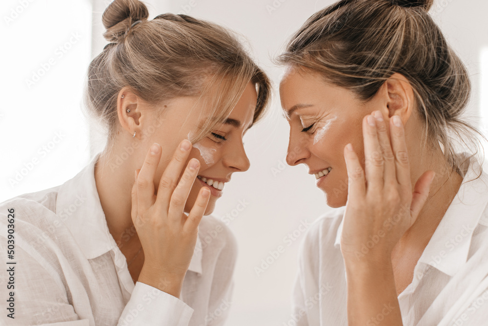 Close-up of two smiling caucasian ladies of different ages with their eyes closed and smeared with cream on white background. Blondes with tied hair in bun wear shirts. Cosmetic products, concept Close-up of two smiling caucasian ladies of different ages with their eyes closed and smeared with cream on white background. Blondes with tied hair in bun wear shirts. Cosmetic products, concept