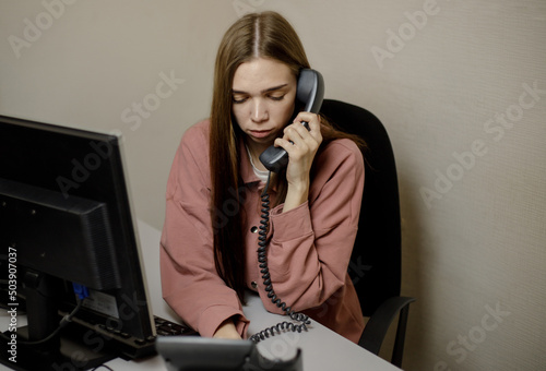 A young girl, an employee in the office is sitting at an office desk, talking on the phone, disappointed, hears bad news, sad, thoughtful, feels unhappy.