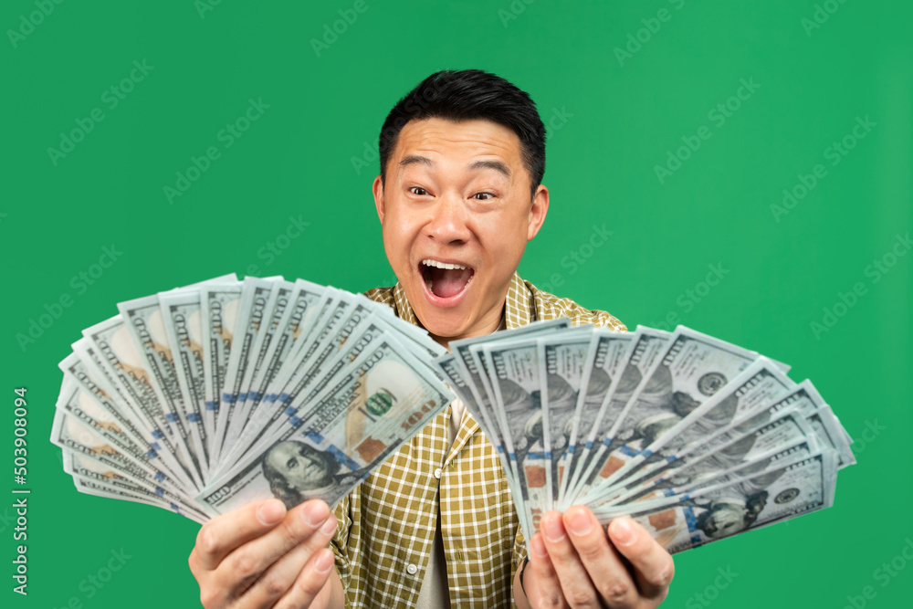 Overjoyed rich mature asian man holding two fans of money, celebrating big financial luck over green background Overjoyed rich mature asian man holding two fans of money, celebrating big financial luck over green background