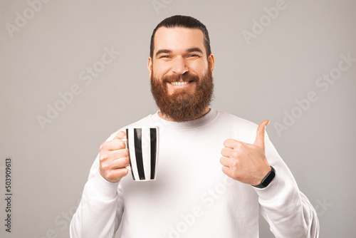 Cheerful young man is holding black and white cup while showing thumb up. Studio shot over grey background. © Vulp
