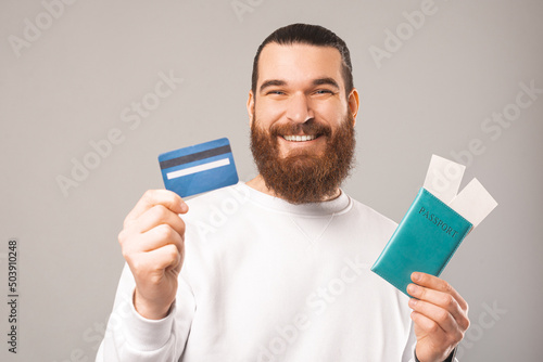 Young smiling bearded hipster man is holding a passport and a credit card. Studio shot over grey background.