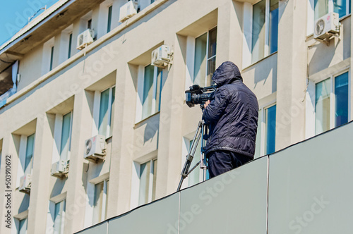 A videographer filming the Victory Day parade from the roof of a building. Professional tripod and video camera. Black jacket and hood. Sunny day. Bottom view. Unknown person.
