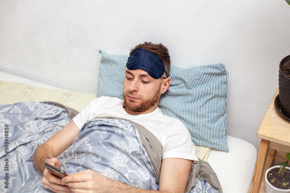 young man lying in bed in the morning wearing a sleep mask and checking social media young man lying in bed in the morning wearing a sleep mask and checking social media