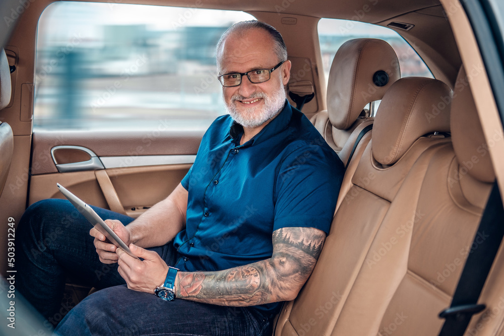 Obraz Portrait of tattooed glad old man executive with tablet sitting inside of car. fototapeta, plakat