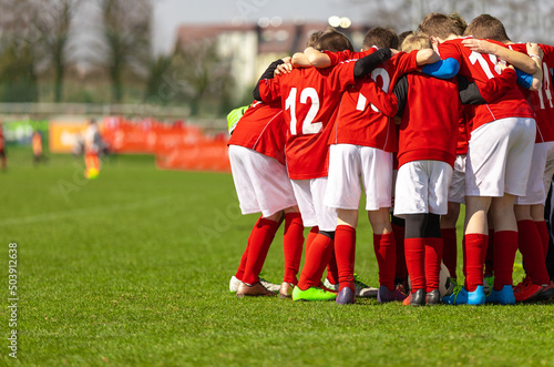 Kids soccer team with coach in group huddle before the match. Elementary age children are listening together to coach motivational speech. Boys in red football shirts