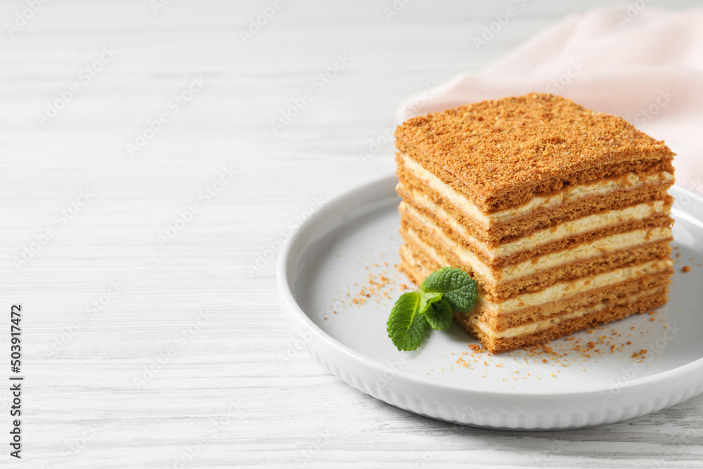 Slice of delicious layered honey cake with mint served on white wooden table. Space for text Slice of delicious layered honey cake with mint served on white wooden table. Space for text