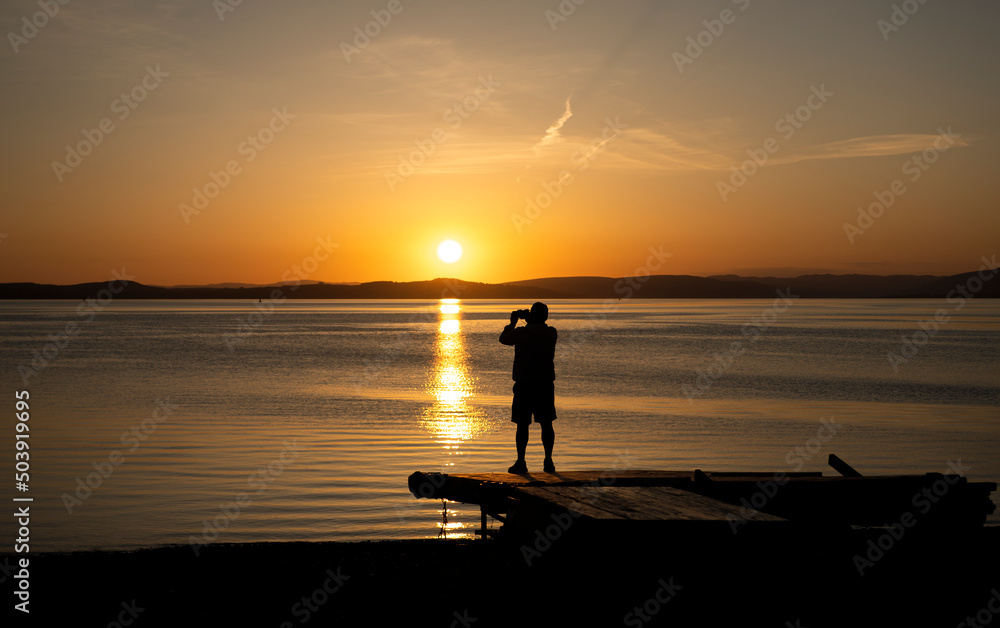 Silhouette of unknown person standing on beach platform with camera taking a picture of beautiful evening sunset.