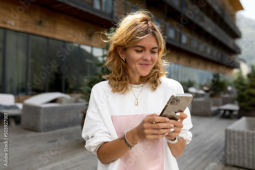 Happy smiling girl is chatting online with friends on smartphone on wooden terrace in. sunlight with happy smile. Pretty girl spend free time outside on the nature