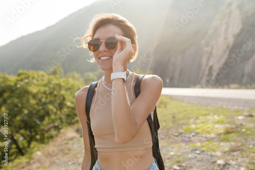 Outdoor portrait of charming stylish european girl with light hair wearing sunglasses and beige t-shirt with backpack is traveling among mountains in sunlight in sunny day