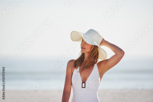 Fotografie A smiling young woman in a white swimsuit holding her hat and enjoying a day at the beach