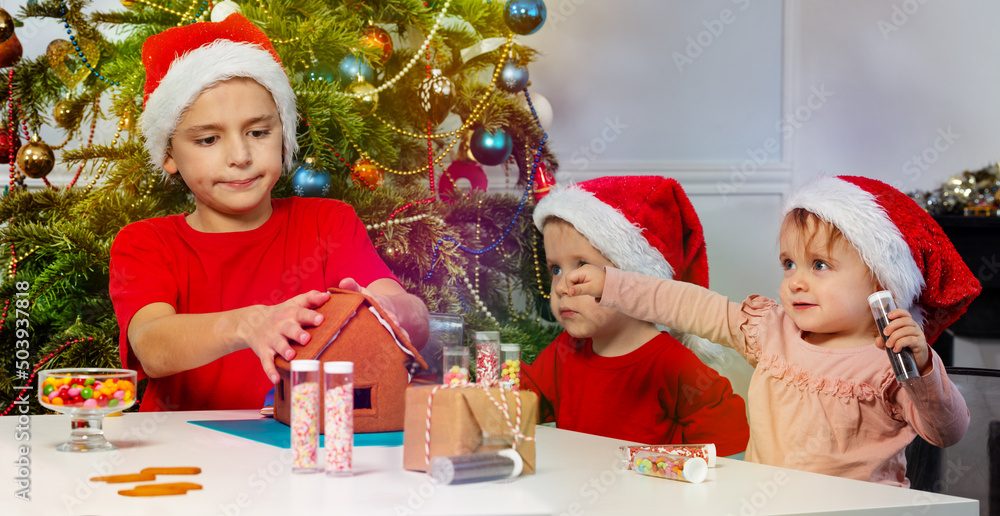 Three kids decorate gingerbread house together in Santa hats