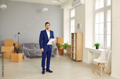 Professional realtor or real estate agent during tour about new apartment or house for sale. Happy young man in suit standing in modern living room or office interior. Real estate business concept