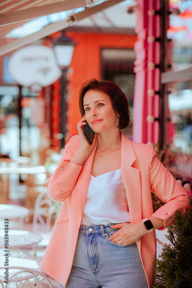 Woman in pink jacket standing at caffe terrace and talking to the cell phone.