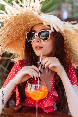 Beautiful girl in sunglasses, straw hat and red polka-dot dress sitting on the summer terrace