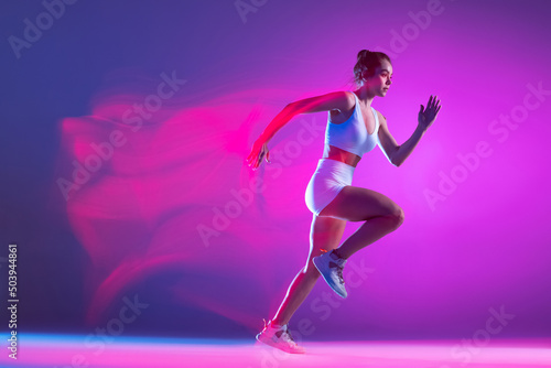 Professional female athlete, runner training isolated on blue studio background in mixed pink neon light. Healthy lifestyle, sport, motion and action concept.