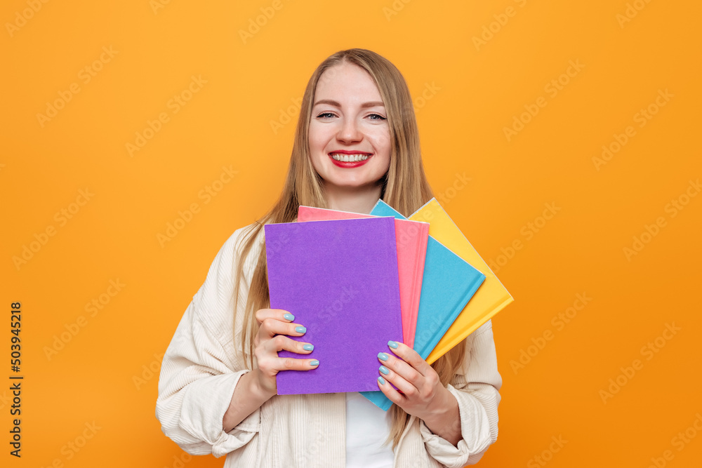 Caucasian blonde girl student in beige jacket holds four books in multi-colored covers smiling isolated on orange background in studio. English language school, education concept Caucasian blonde girl student in beige jacket holds four books in multi-colored covers smiling isolated on orange background in studio. English language school, education concept