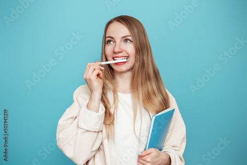 blonde girl student thoughtful, daydreaming and bit her handle with her teeth isolated on a blue studio background. Education concept