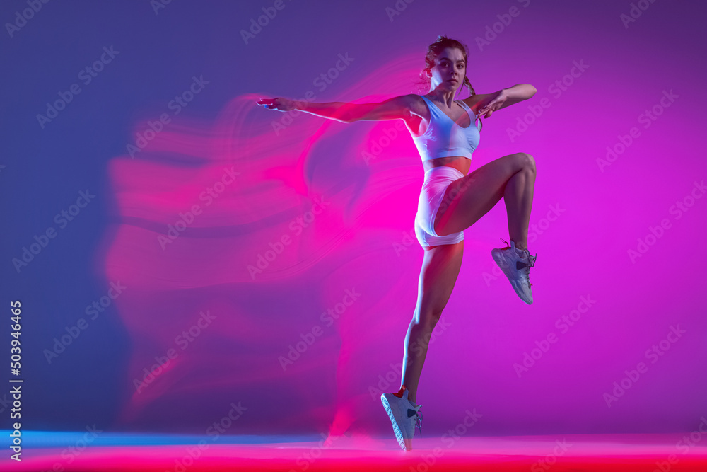 Young female athlete, runner training isolated on blue studio background in mixed pink neon light. Healthy lifestyle, sport, motion and action concept.