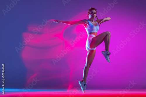 Fototapeta Naklejka Na Ścianę i Meble -  Young female athlete, runner training isolated on blue studio background in mixed pink neon light. Healthy lifestyle, sport, motion and action concept.