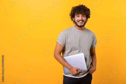 Photographie Cheerful Indian young man holding laptop standing isolated on yellow, looking at camera with happy smile