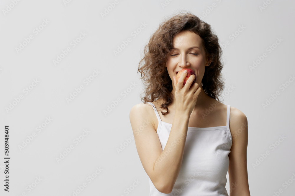 Enjoyed cheerful lovely curly beautiful woman in basic white t-shirt enjoy fresh apple fruit posing isolated on over white background. Natural Eco-friendly products concept. Copy space Enjoyed cheerful lovely curly beautiful woman in basic white t-shirt enjoy fresh apple fruit posing isolated on over white background. Natural Eco-friendly products concept. Copy space