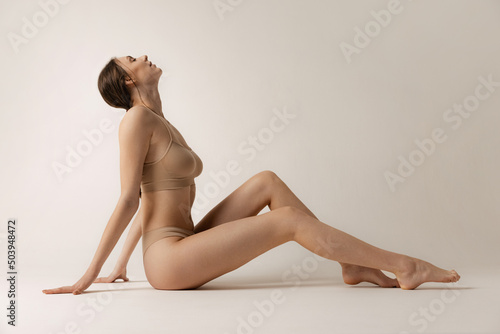 Side view portrait of young slim woman sitting on floor, posing in beige underwear isolated over grey studio background. Depilation, epilation concept