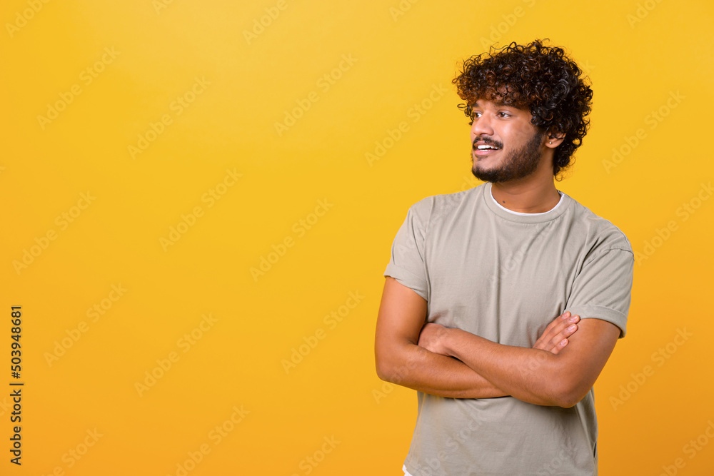 Portrait of happy satisfied handsome young Indian man in t-shirt standing with crossed arms and looking away. Indoor studio shot on yellow background Portrait of happy satisfied handsome young Indian man in t-shirt standing with crossed arms and looking away. Indoor studio shot on yellow background