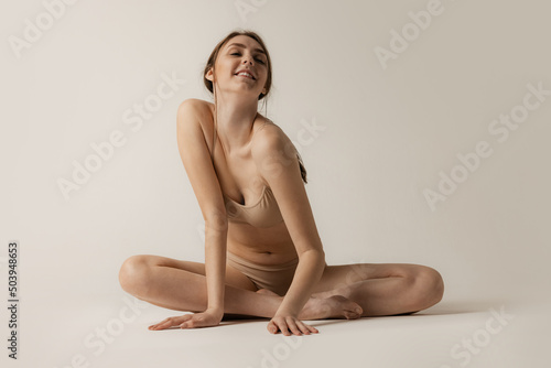 Portrait of young cheerful girl sitting on floor, smilig, posing in beige underwear isolated over grey studio background