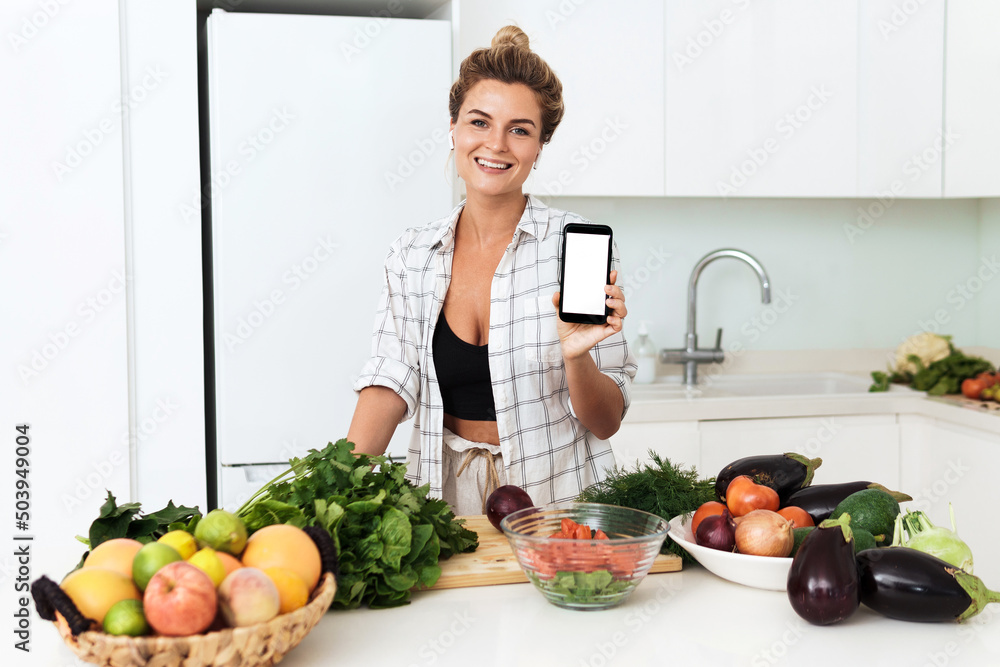 Woman is holding smartphone with blank screen for your design during cooking in modern kitchen