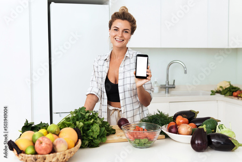 Woman is holding smartphone with blank screen for your design during cooking in modern kitchen