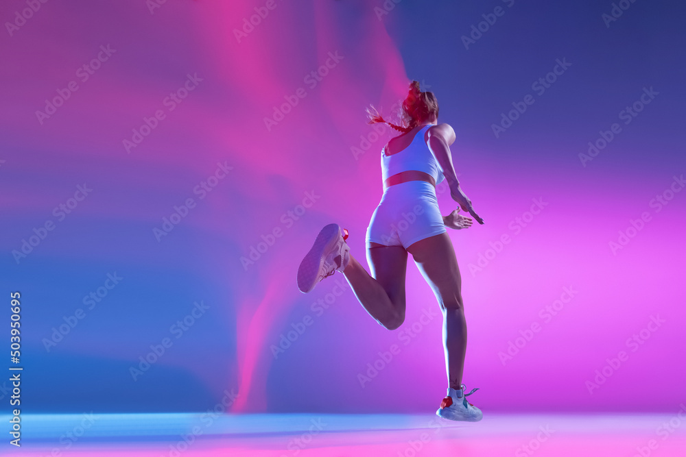 Back view. Professional female athlete running away isolated on blue studio background in mixed pink neon light. Healthy lifestyle, sport, motion and action concept.