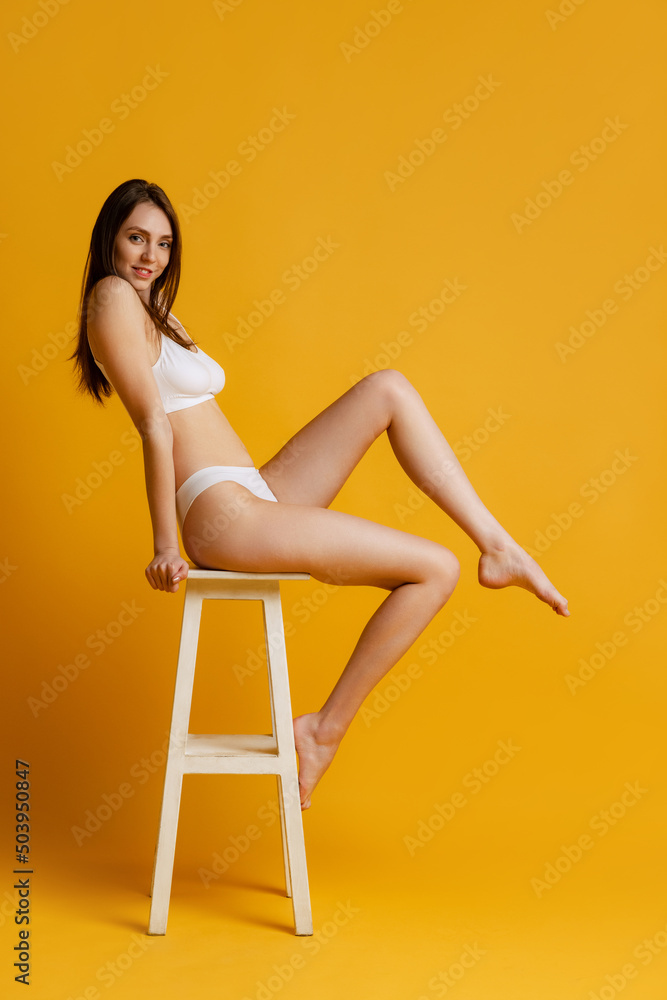 Naklejka premium One tender young girl sitting on chair, posing in white underwear isolated over yellow studio background. Smooth legs