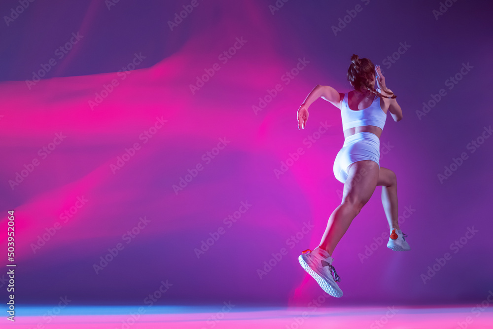 back view. Professional female athlete, runner training isolated on blue studio background in mixed pink neon light.
