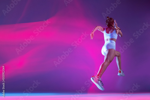 back view. Professional female athlete, runner training isolated on blue studio background in mixed pink neon light. © master1305