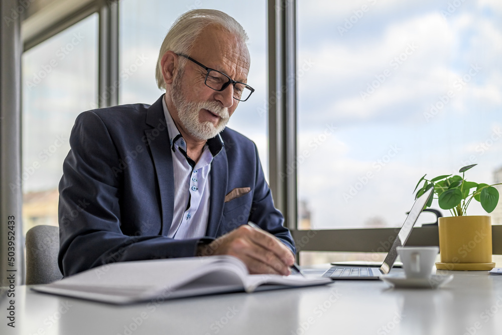 Serious businessman writing business plan by laptop at desk in corporate office Serious businessman writing business plan by laptop at desk in corporate office