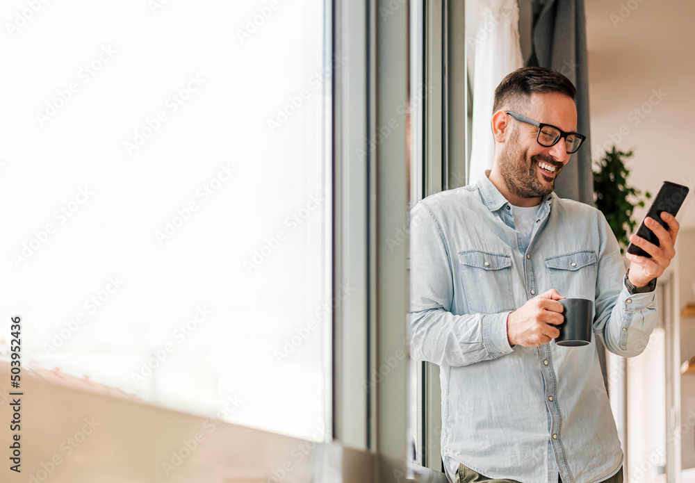 Smiling man businessman in casuals standing in office next to window using mobile phone drinking coffee holding coffee cup Small business entrepreneur looking at smart phone while taking coffee break