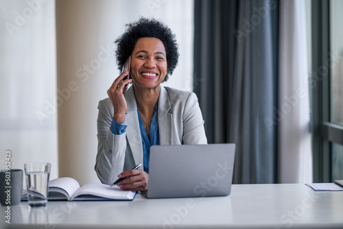 Portrait of happy entrepreneur businesswoman talking on cellphone while using laptop at desk.