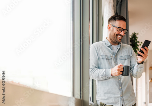 Smiling man businessman in casuals standing in office next to window using mobile phone drinking coffee holding coffee cup Small business entrepreneur looking at smart phone while taking coffee break