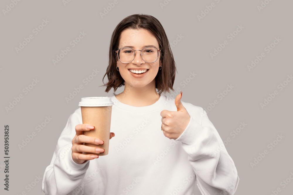 Cheerful woman is holding a disposable paper cup while showing thumb up. Studio shot over grey background. Cheerful woman is holding a disposable paper cup while showing thumb up. Studio shot over grey background.