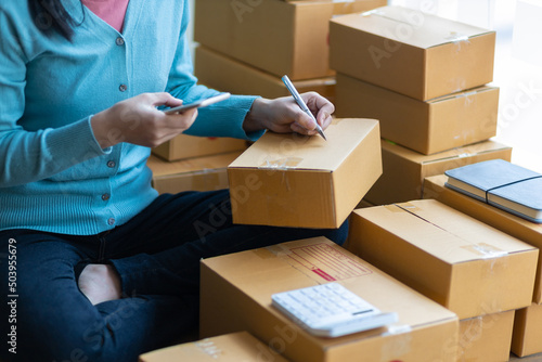 Shipping shopping online, startup small business owner writing address on cardboard box at workplace. Freelance Asian woman small business entrepreneur SME working with box at home.