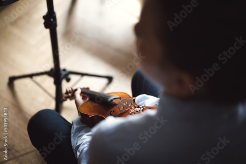 Teenage violin player sitting indoors with the instrument in his hands, over the shoulder view
