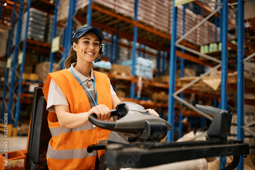 Happy female worker driving pallet jack while working at distribution warehouse.