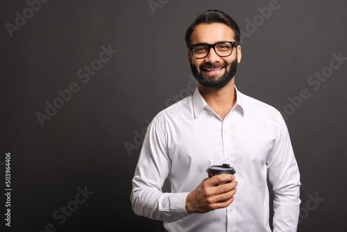 Handsome confident bearded Indian man in formal wear stands with paper cup of coffee to-go isolated on black, smiling male office employee in glasses take a coffee break © Vadim Pastuh
