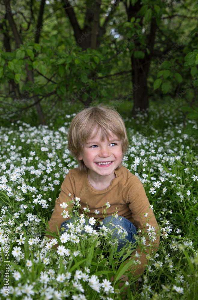 cute cheerful boy sits among many small flowers and bright green grass in forest. Walk in nature in the park. Spring flowering. walking in pleasure outdoors. joyful childhood. Earth Day