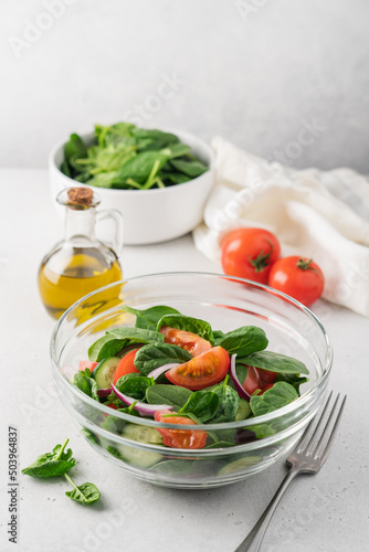 Fresh salad in glass transparent bowl on white background. Spinach, tomato, cucumber, onion fresh vegetables for vegan, healthy diet