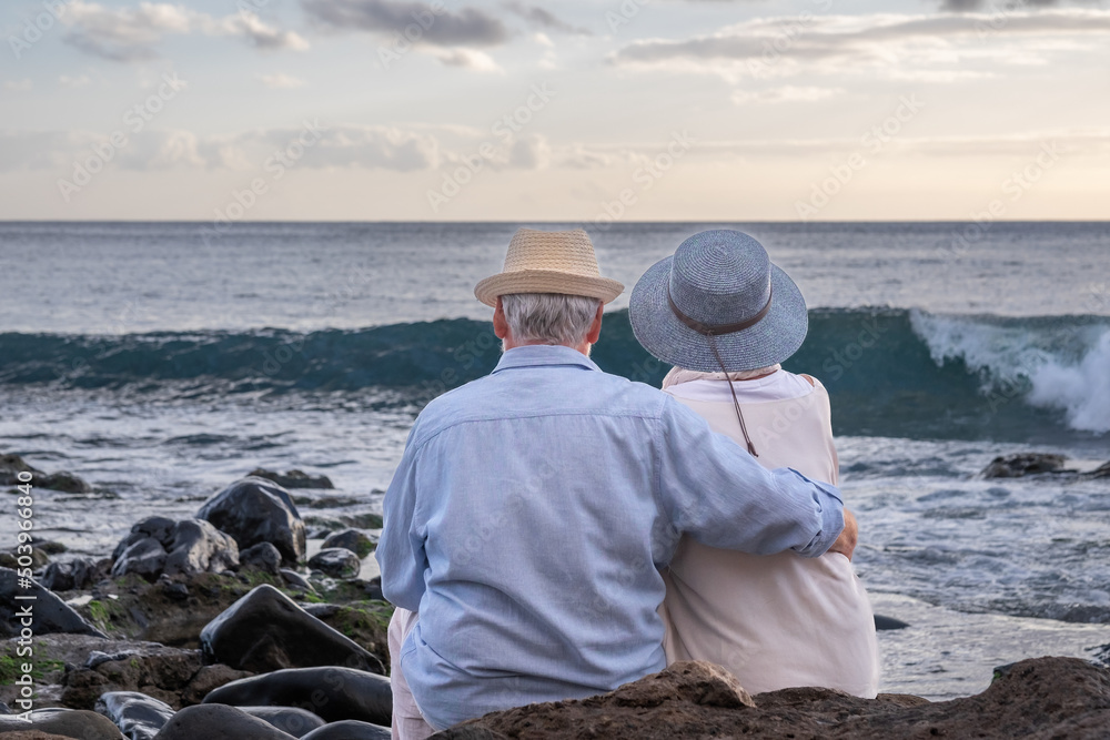 Rear view of relaxed caucasian senior couple sitting on the pebble beach at sunset light admiring horizon over water. Two gray haired elderly people hugging with love