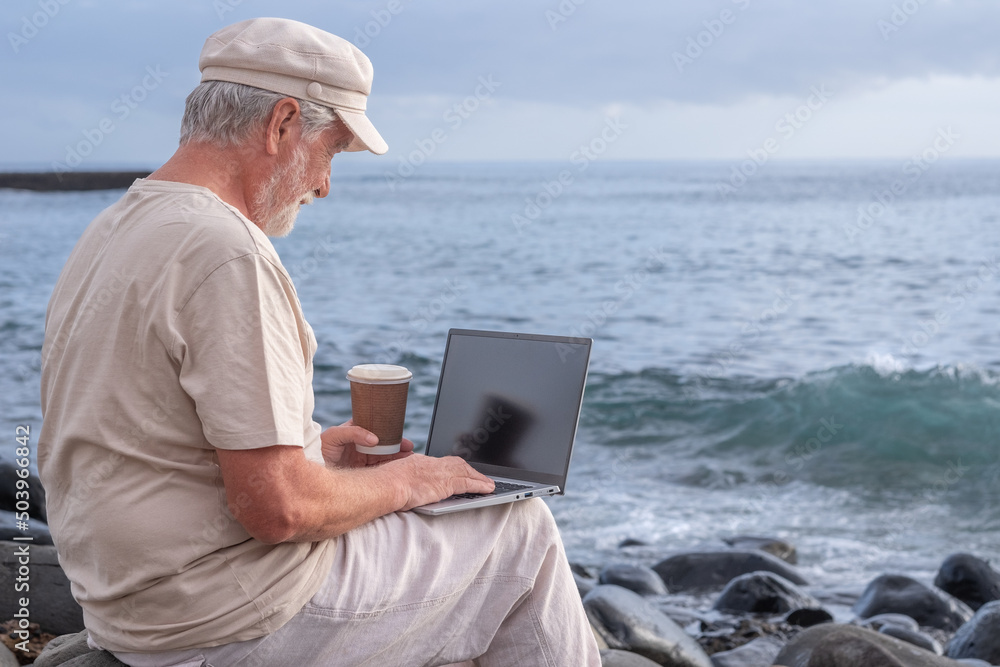 Senior active man sitting on the beach using laptop. Elderly bearded male holding a coffee cup enjoying sunset at sea, horizon over water